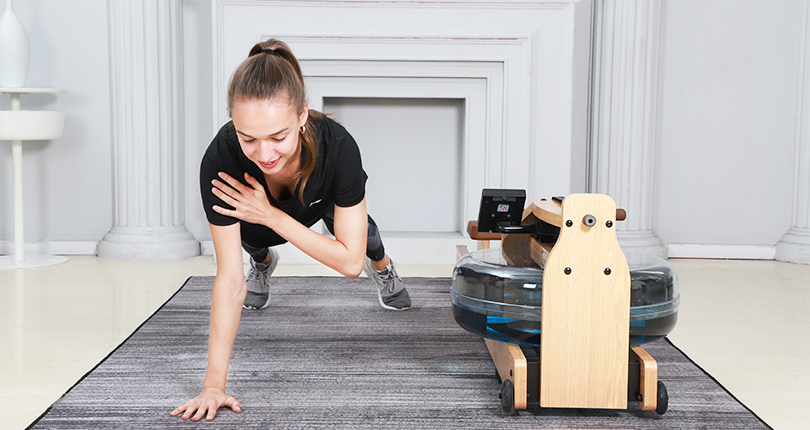a woman doing single arm plank beside a blue-dyed rowing machine in a room