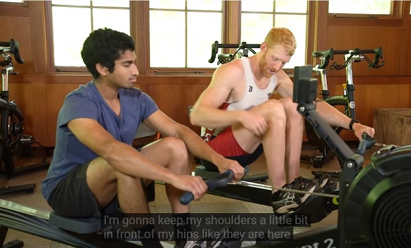 One man with black hair and another man with yellow hair keep their shoulders  in front of hips when doing the recovery motion of indoor rowing.